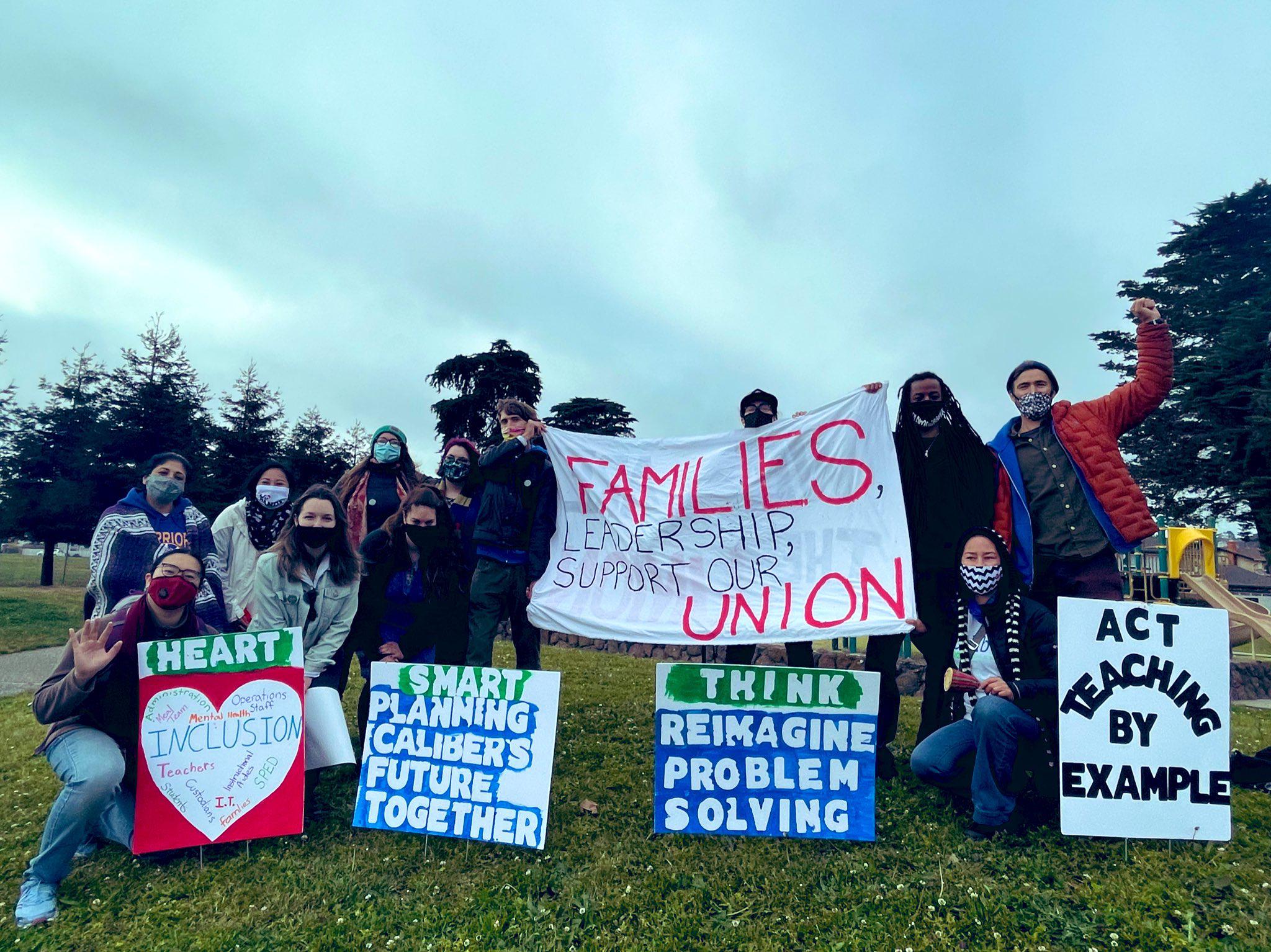 Members of the Caliber Workers Union gathered on grass holding a variety of signs and a banner in support of teachers and their union.